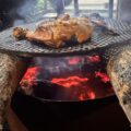 Chicken meat grilling on a metal grate over glowing red coals, supported by logs, with smoke rising from the fire.